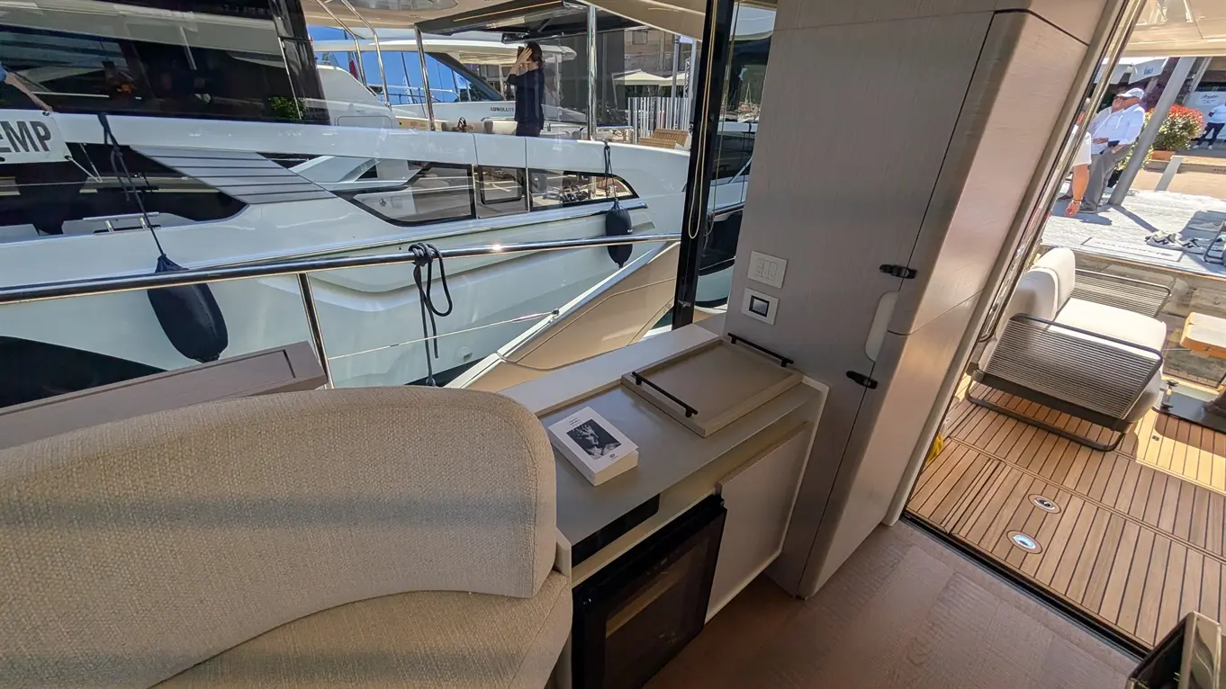 Interior view of a modern yacht, featuring a curved sofa, a desk with a book, and large windows overlooking docked boats.